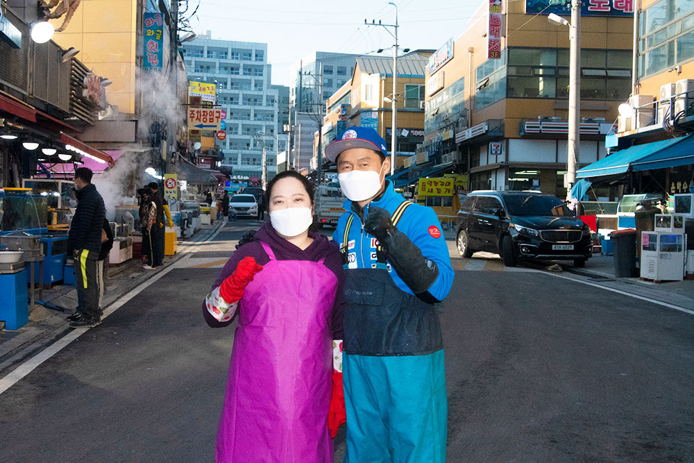 하남수산물시장내 메인 도로위에 왼쪽과 오른쪽에는 도매매장들이 있으며 은성수산 매장 앞 도로중앙에 은성수산 대표 부부가 남자는 파란색 도롱이를 입고 파란색 야구모자와 흰색 마스크 검정색 고무장갑을 끼고 있으며 왼쪽 부인은 분홍색 앞치마에 빨간색 고무장갑을 끼고 웃는 사진입니다